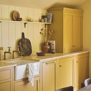 Shaker style wooden kitchen units painted in yellow paint with wood panel splashback, butler sink and open shelf