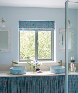 Blue bathroom with double floral basins, blue walls, blue fabric skirt, blind, pale blue walls,