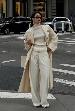 a photo showing the best summer-to-fall transition items with a woman standing in the street wearing a white trench coat over a white high-neck top with white trousers, a white belt, white bag, and black shoes