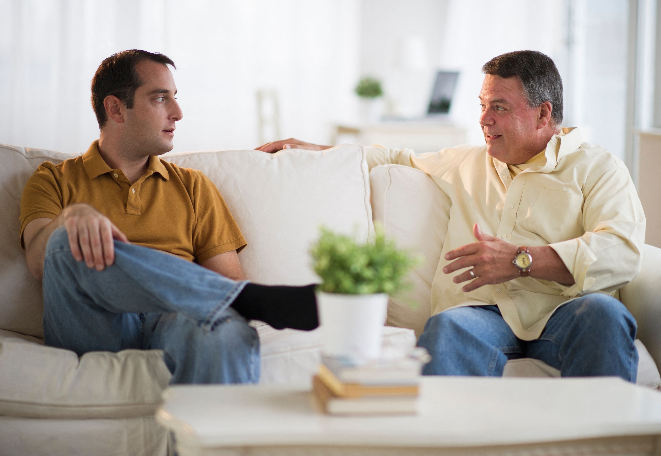 A father and his adult son sit on the couch talking.