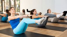 Three women in a group Pilates class practice the Hundred exercise on black mats. They are seated, with their knees bent and legs elevated in the air, and their torsos upright, arms parallel to the floor. 
