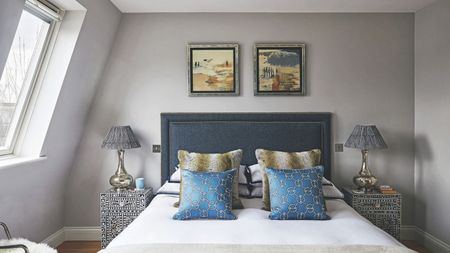A grey bedroom with a double bed with blue headboard, white bedsheets and blue, white and green patterned cushions. On either side of the bed are black and white patterned bedside tables, and above are two framed artworks in silver framed. To the left is a slanted window.
