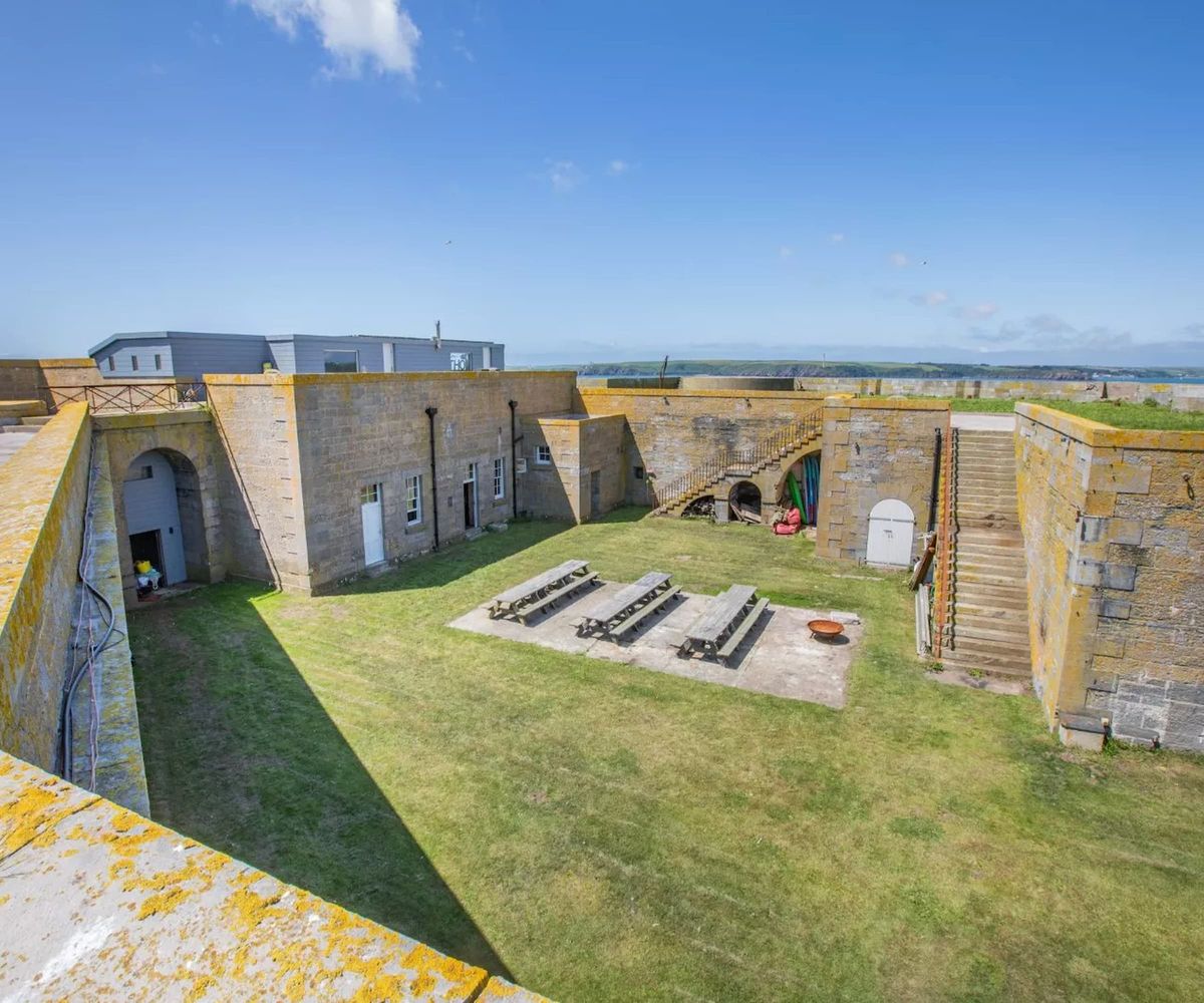 A courtyard with three large tables and a grass garden surrounded by staircases leading to rooftop balconies