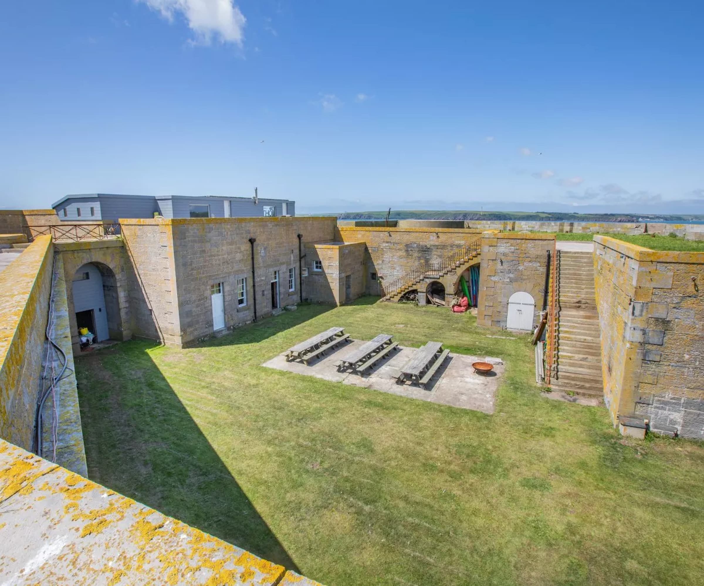 A courtyard with three large tables and a grass garden surrounded by staircases leading to rooftop balconies