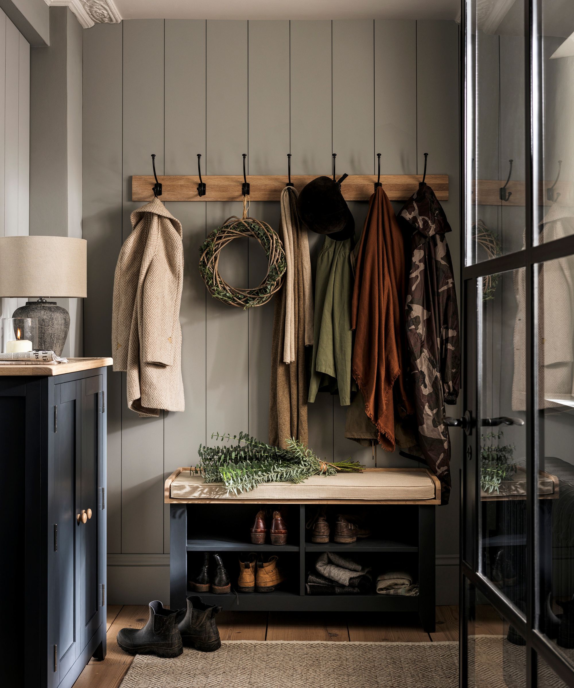 A rustic entryway with wall mounted coat hooks above a blue painted shoe storage bench, topped with a cream cushion.