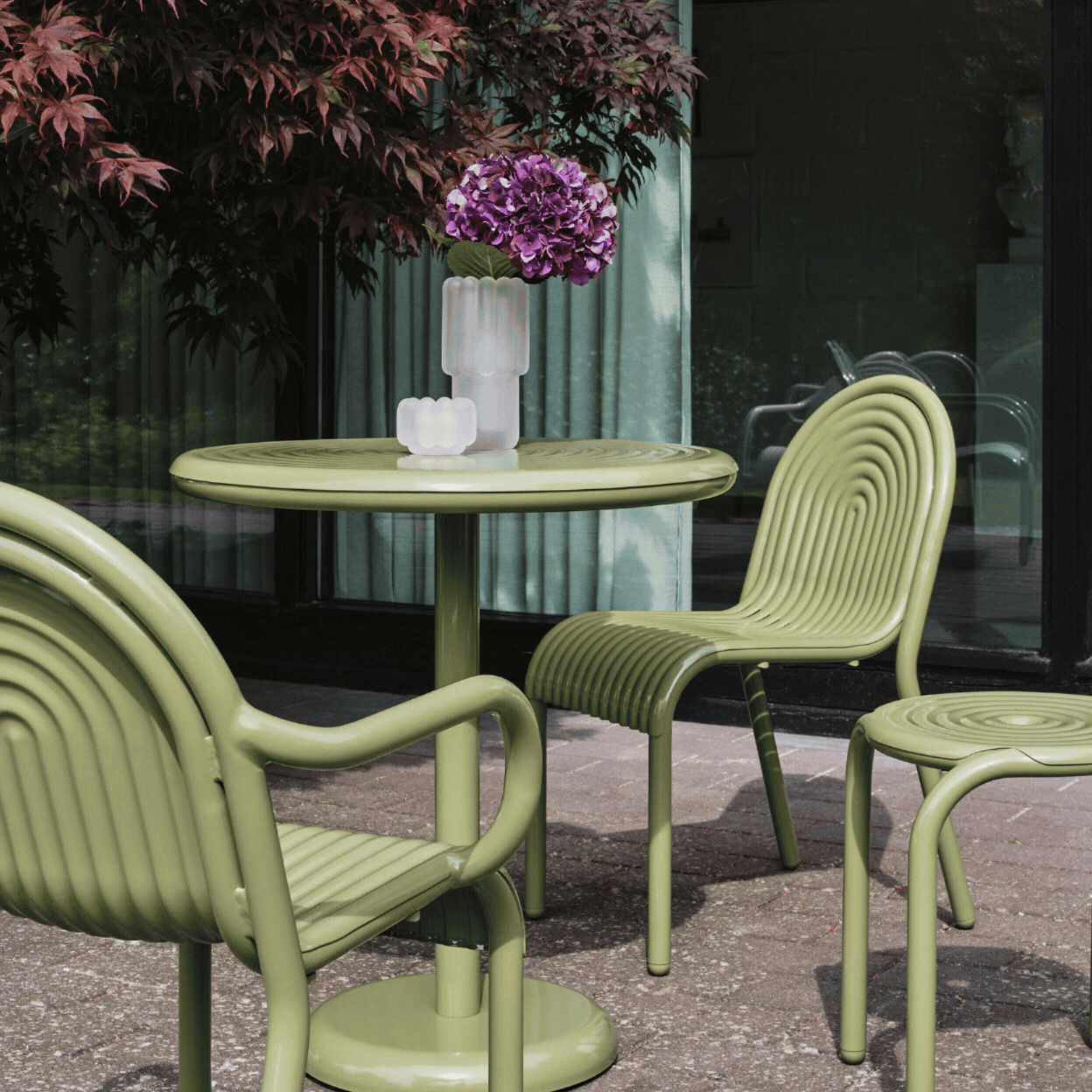 Groove Outdoor Small Round Table in Moss Weather-Resistant Aluminium showcased in a rotating gif in a sun-filled outdoor patio, topped with glass vases and purple flowers, and as a product against a pale green background.