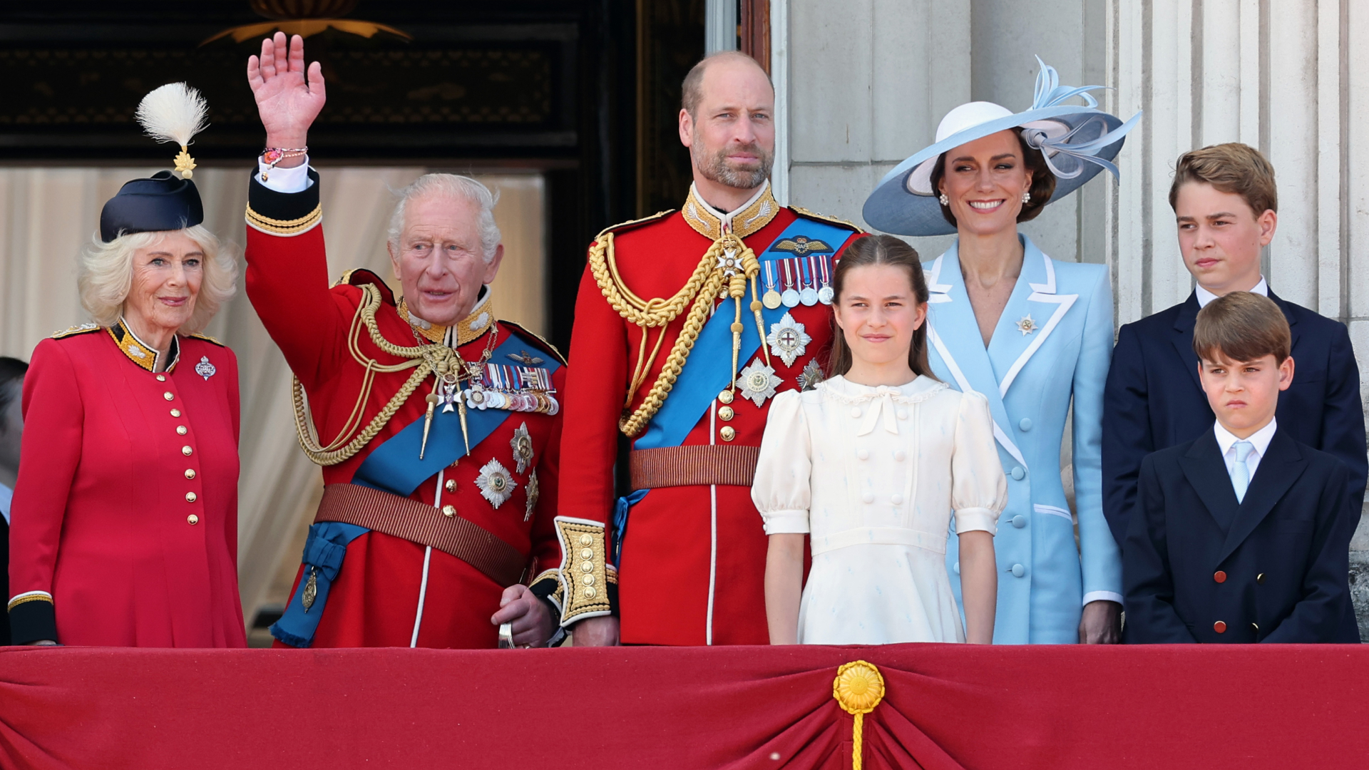 King Charles waves from the Buckingham Palace balcony while wearing a red military uniform