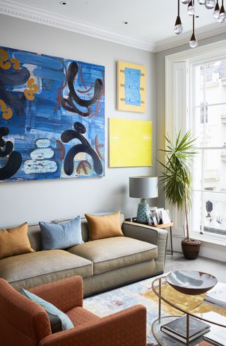 Colorful small living room with statement art in bright yellows and blues, an orange armchair and round glass coffee table.