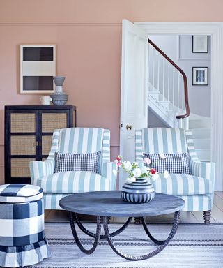 Pink painted living room with two blue and white striped armchairs, black round coffee table, black and white check pouf, black cabinet with rattan doors, looking onto white painted stairway