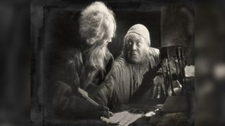 An old man with a long white beard and wild hair signs a parchment with a quill, while a wide-eyed figure in a skullcap leans in beside him, one clawed hand resting on the table next to an hourglass