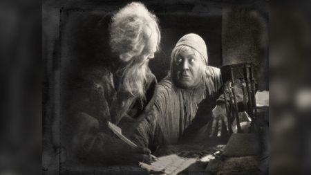 An old man with a long white beard and wild hair signs a parchment with a quill, while a wide-eyed figure in a skullcap leans in beside him, one clawed hand resting on the table next to an hourglass