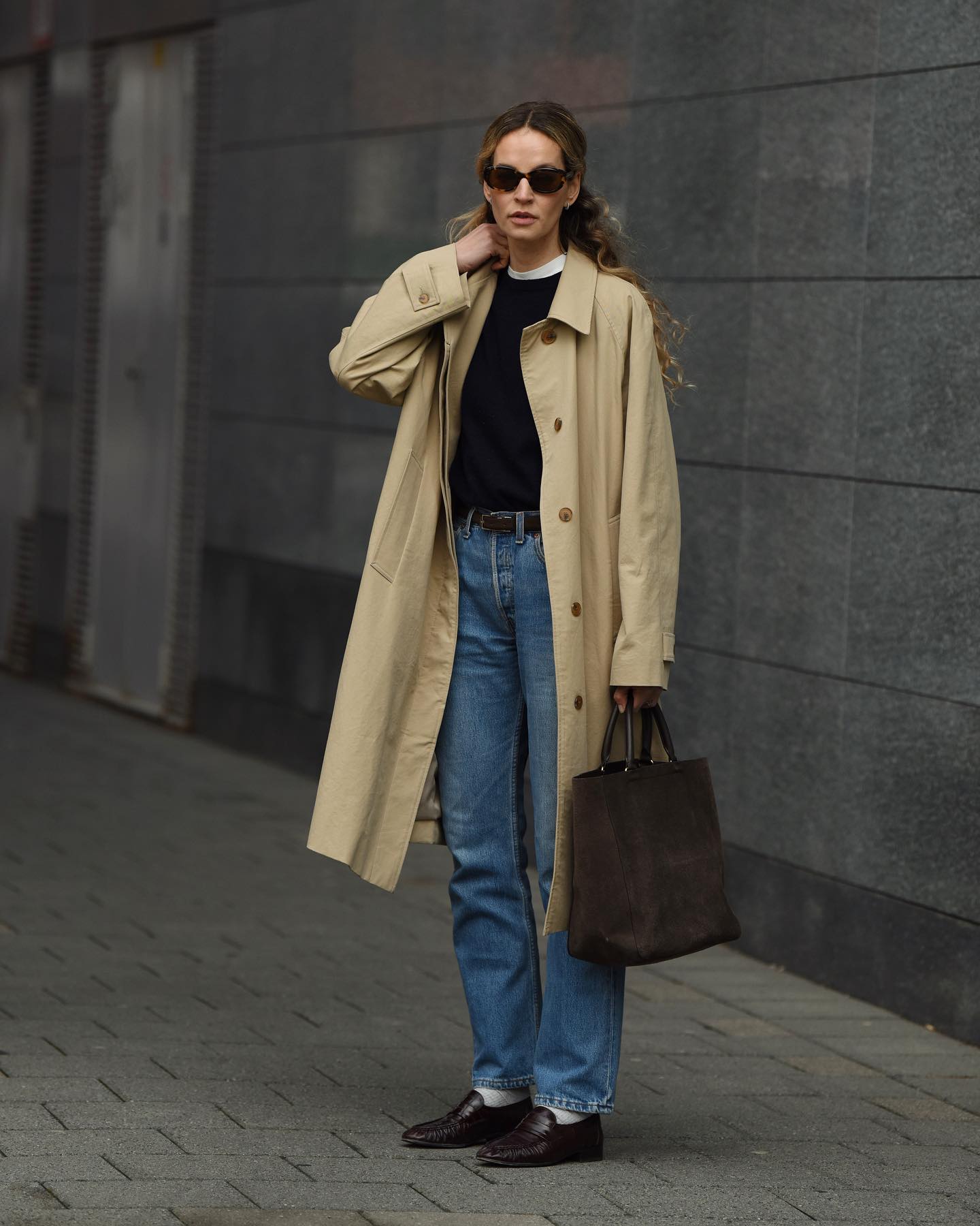 Woman wearing khaki trench coat, black sweater, black sunglasses, belt, blue jeans, loafers, and carrying brown suede handbag