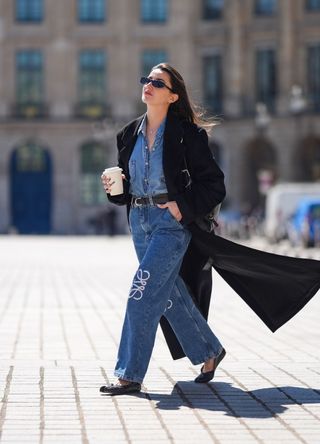 Alexandra Pereira wears sunglasses, a black long trench coat , a blue denim shirt, a belt, blue denim pants / jeans with embroidered logo from Loewe, black leather flat ballerina shoes with separated toes, a black leather Givenchy Voyou bag