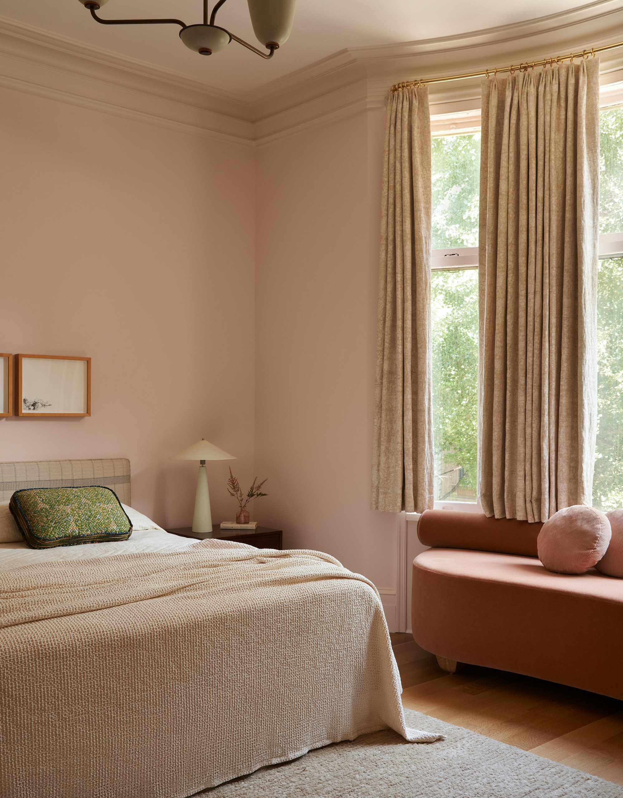 A blush bedroom with a textured blanket, a patterned pillow, a white lamp, and a blush bench by a large window