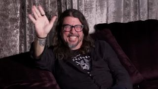 Dave Grohl waving and smiling on a dark couch
