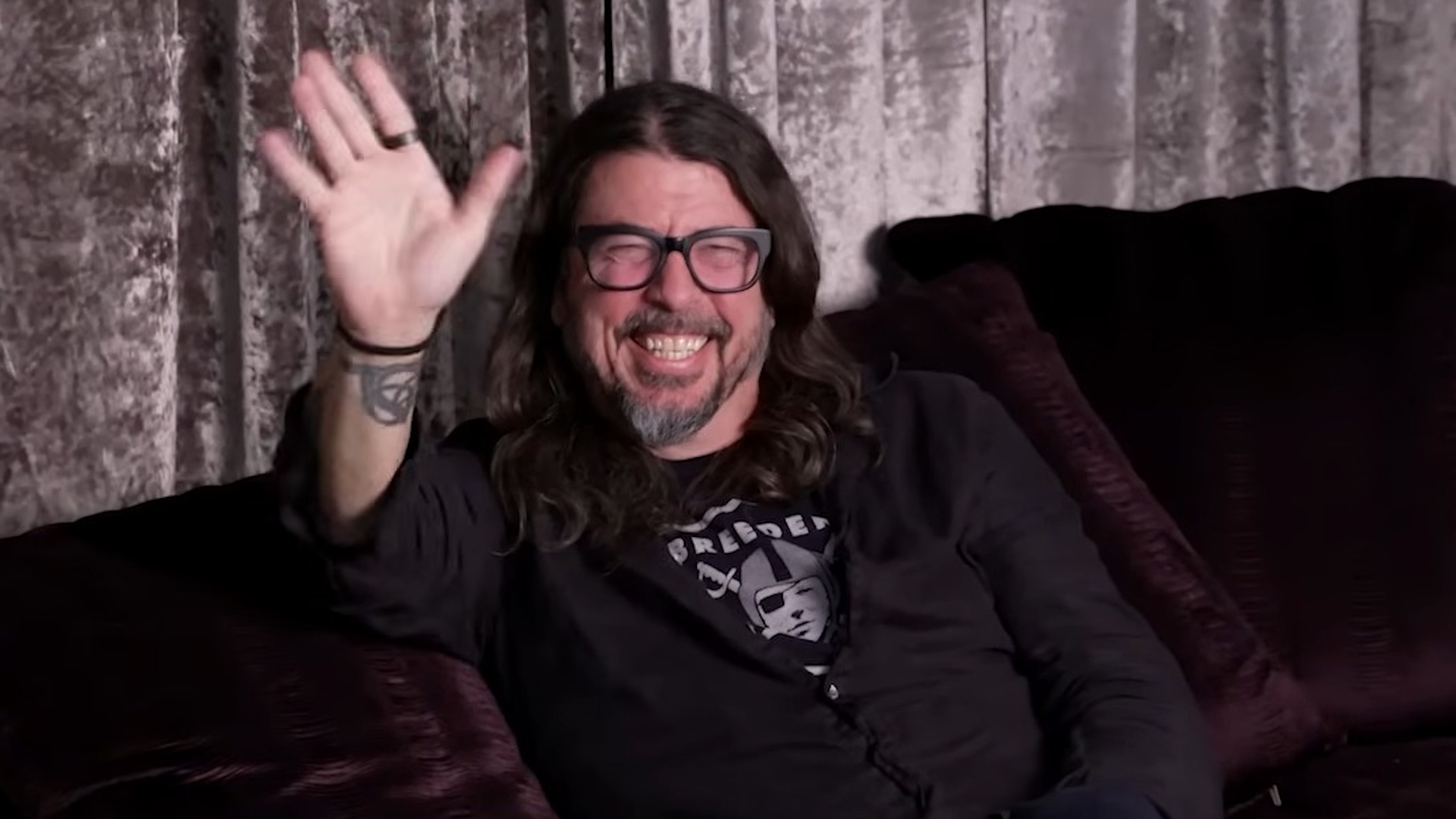 Dave Grohl waving and smiling on a dark couch