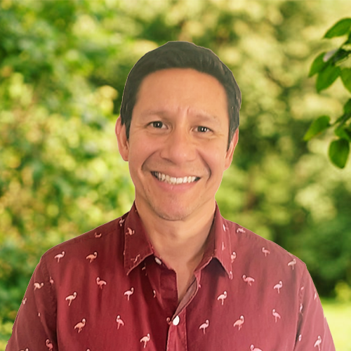 Dr Greg Gomez is a white man wearing a red collared shirt with flamingos on, smiling broadly outside, set against a background of green trees and shrubs
