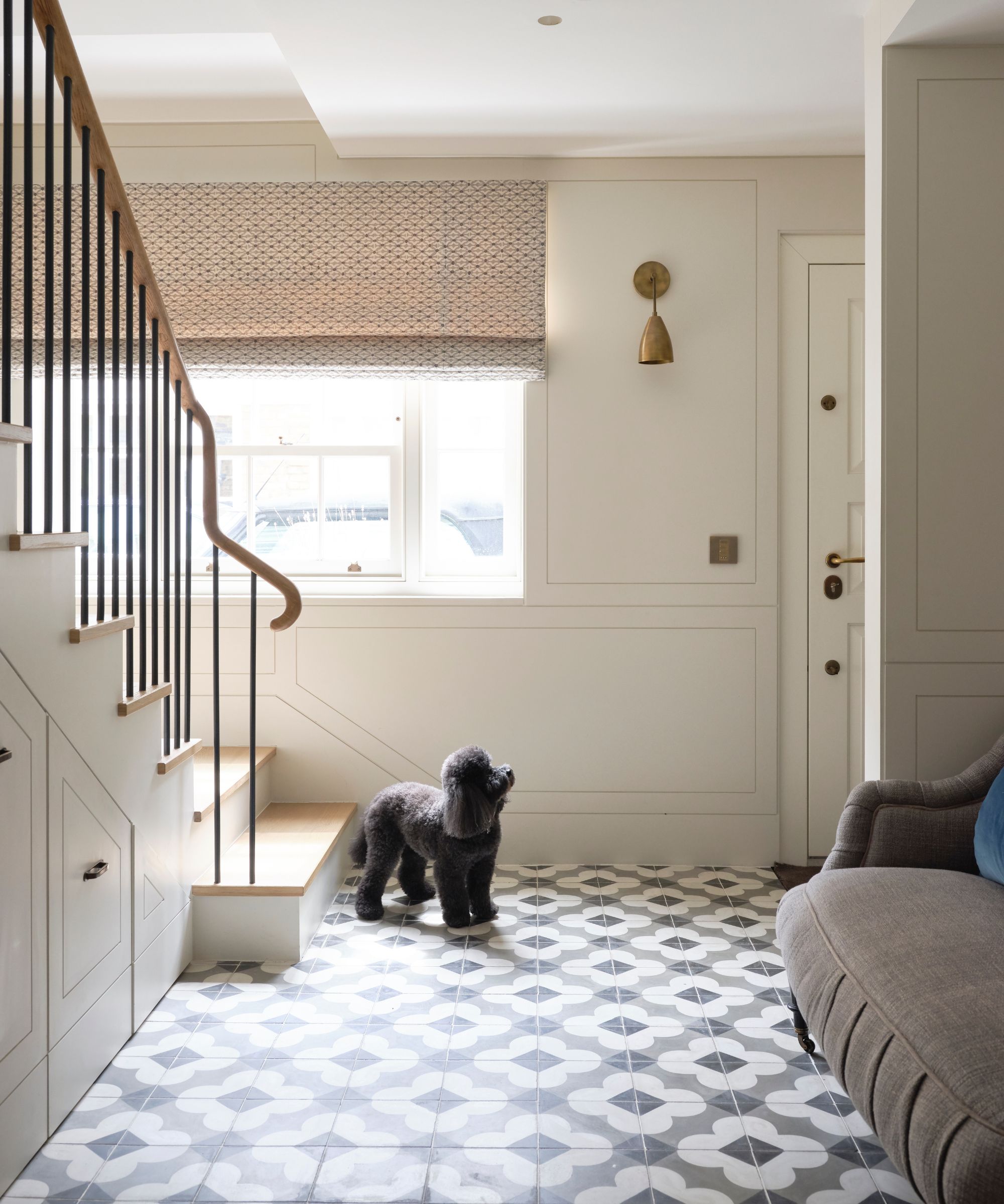 A white entryway with black and white tiles, a white and wood staircase, brown couch, white front door, gold wall light, large window with patterned blind, and a small black poodle dog.