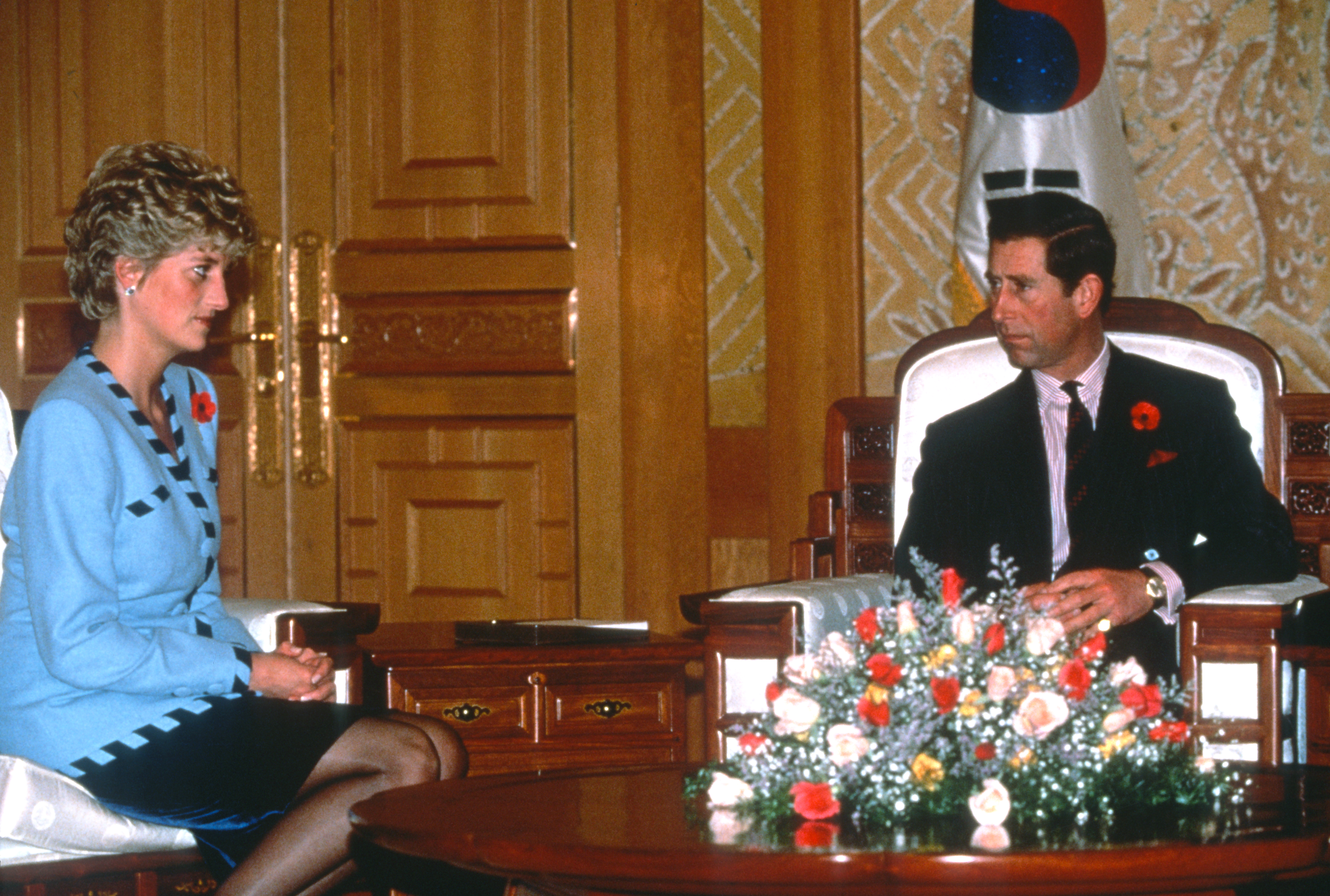 Princess Diana sitting in a blue and black suit giving Prince Charles a dirty look