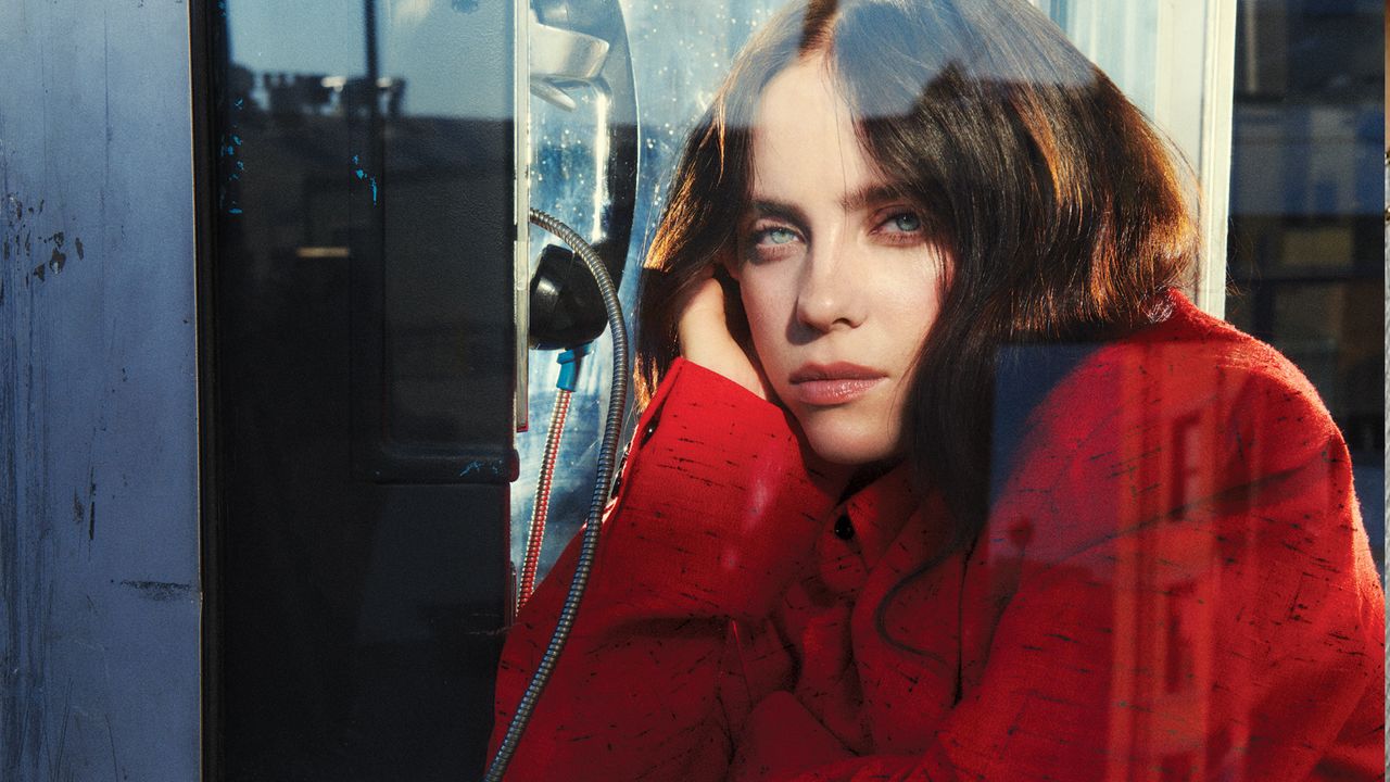 Billie Eilish wearing a red coat posing with her hand on her cheek inside of a phone booth