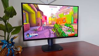 Alienware AW2725Q monitor on woodgrain desk next to plant with Overwatch 2 gameplay on screen featuring Pharah with jade green gun skin.