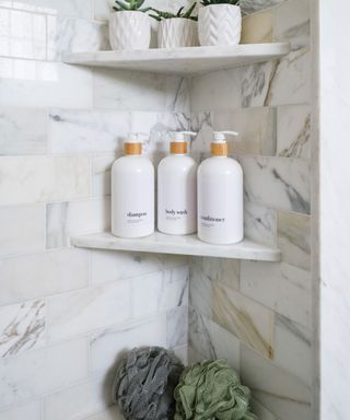 A marble corner of a shower with three corner shelves with plants, white toiletry bottles, and poufs on them