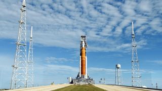 The Artemis 1 satellite  rocket stands gangly  connected  the motorboat  pad with bluish  entity  and clouds above. 