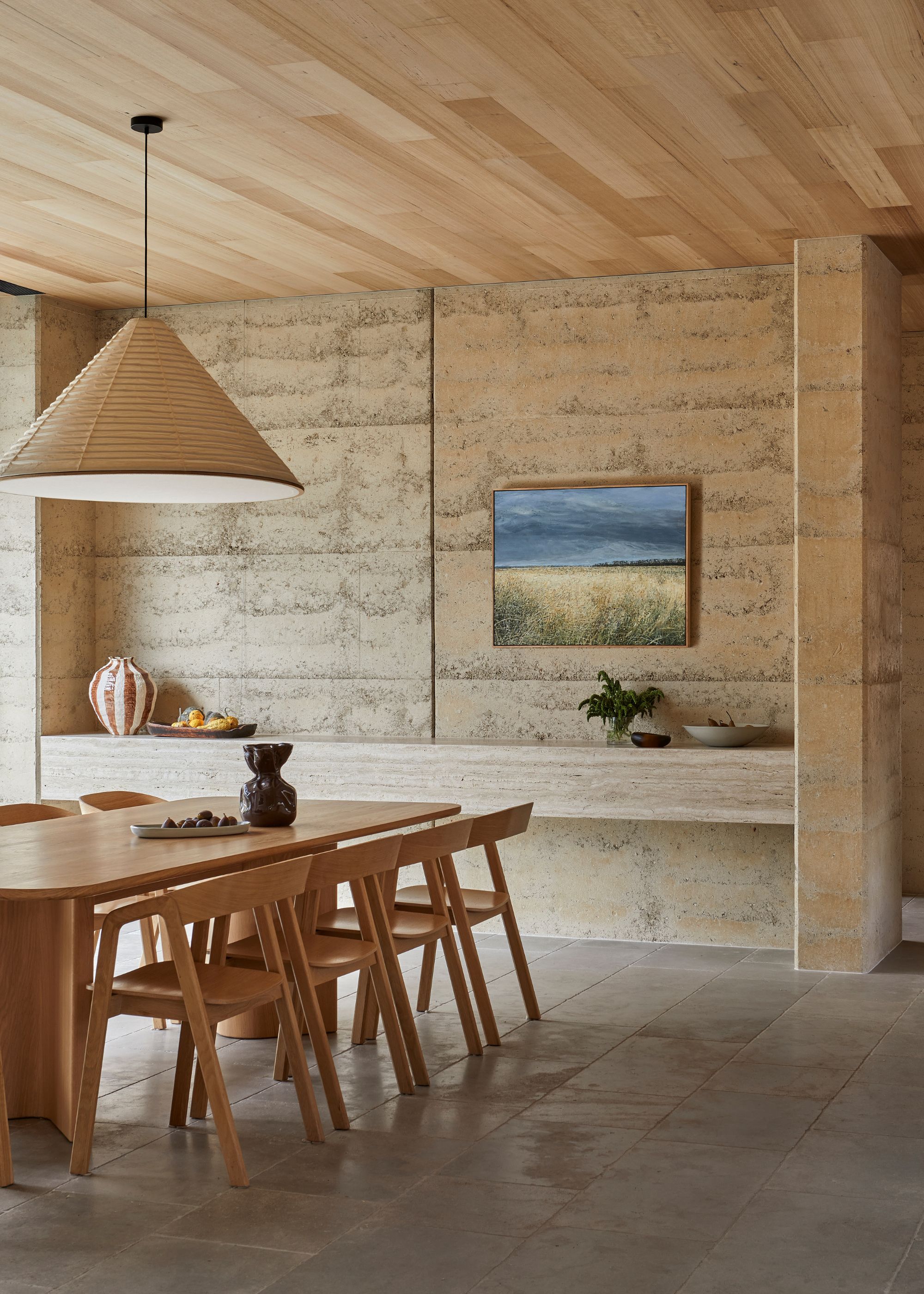 A minimalist dining room with a long shelf, a wood dining set, a pendant lantern, a textured base, and a bowl of fruit