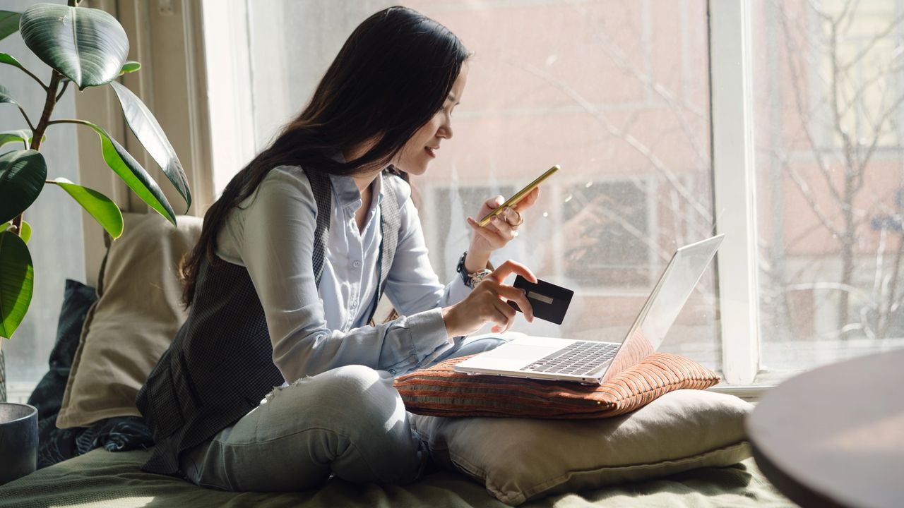 A middle-aged asian woman in blue jeans sitting on the bed in a yoga pose in front of a laptop - stock photo