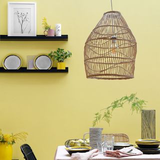 Dining table with pink tablecloth, yellow walls, black shelves and chairs and large wicker lampshade