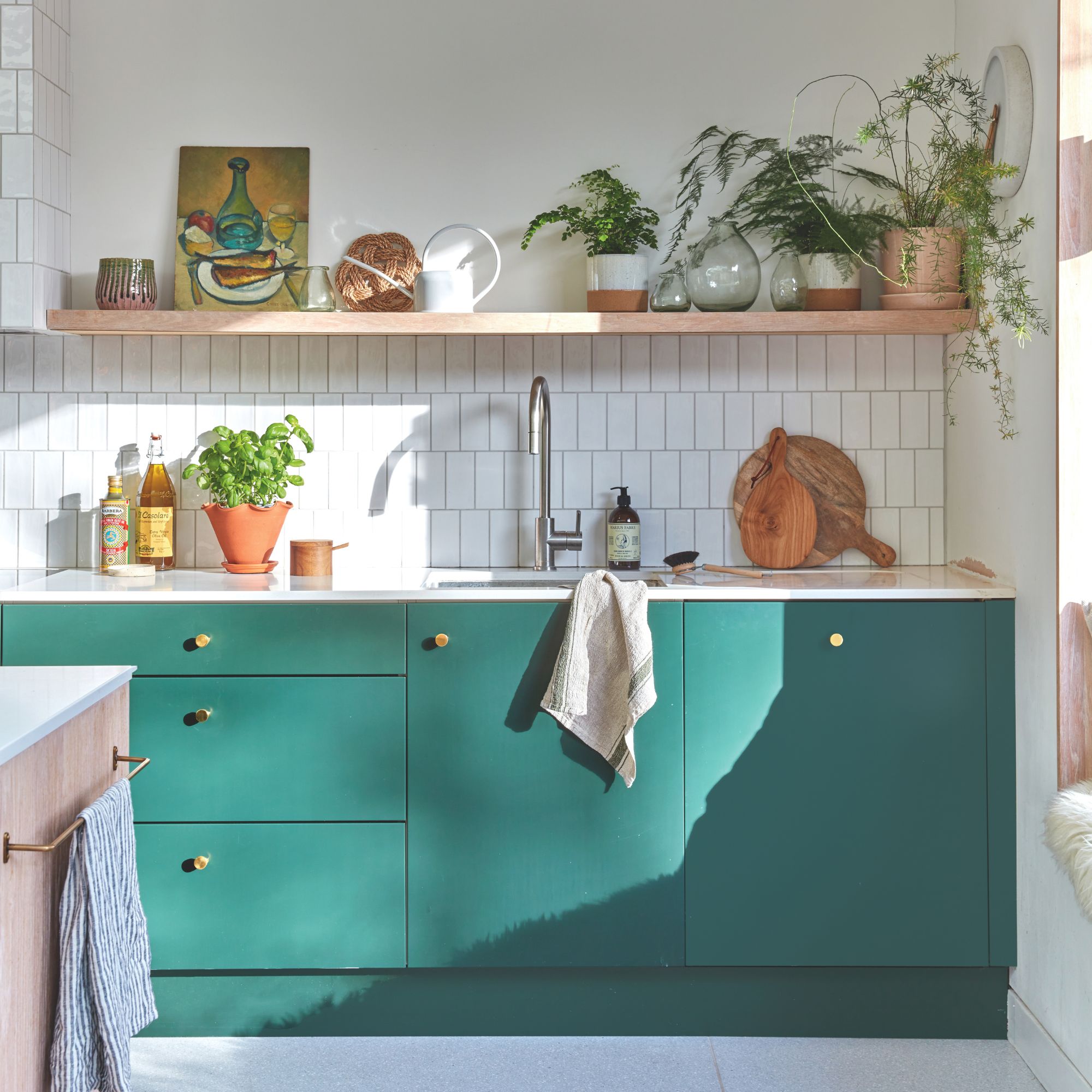 White tiled kitchen with teal painted cabinets underneath a sink and floating shelf