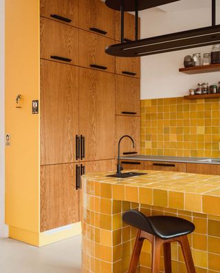 yellow kitchen with yellow tiled counter, island, and backsplash, yellow panel on cabinetry with timber finish, black handles, floating shelves