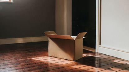 An open cardboard box in the middle of an empty room with wooden floors and gray painted walls