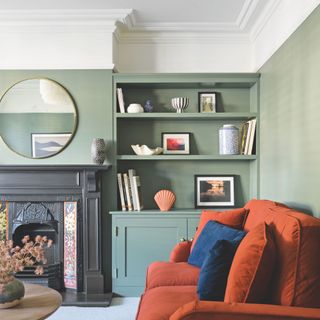 Green living room with shelves on the wall and white coving, with an orange sofa with blue cushions on it