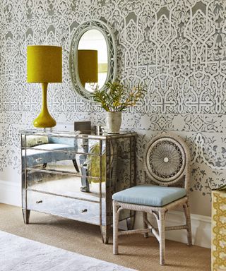 A bedroom furniture idea with mirrored chest of drawers, patterned wallpaper, yellow lamp and rattan chair