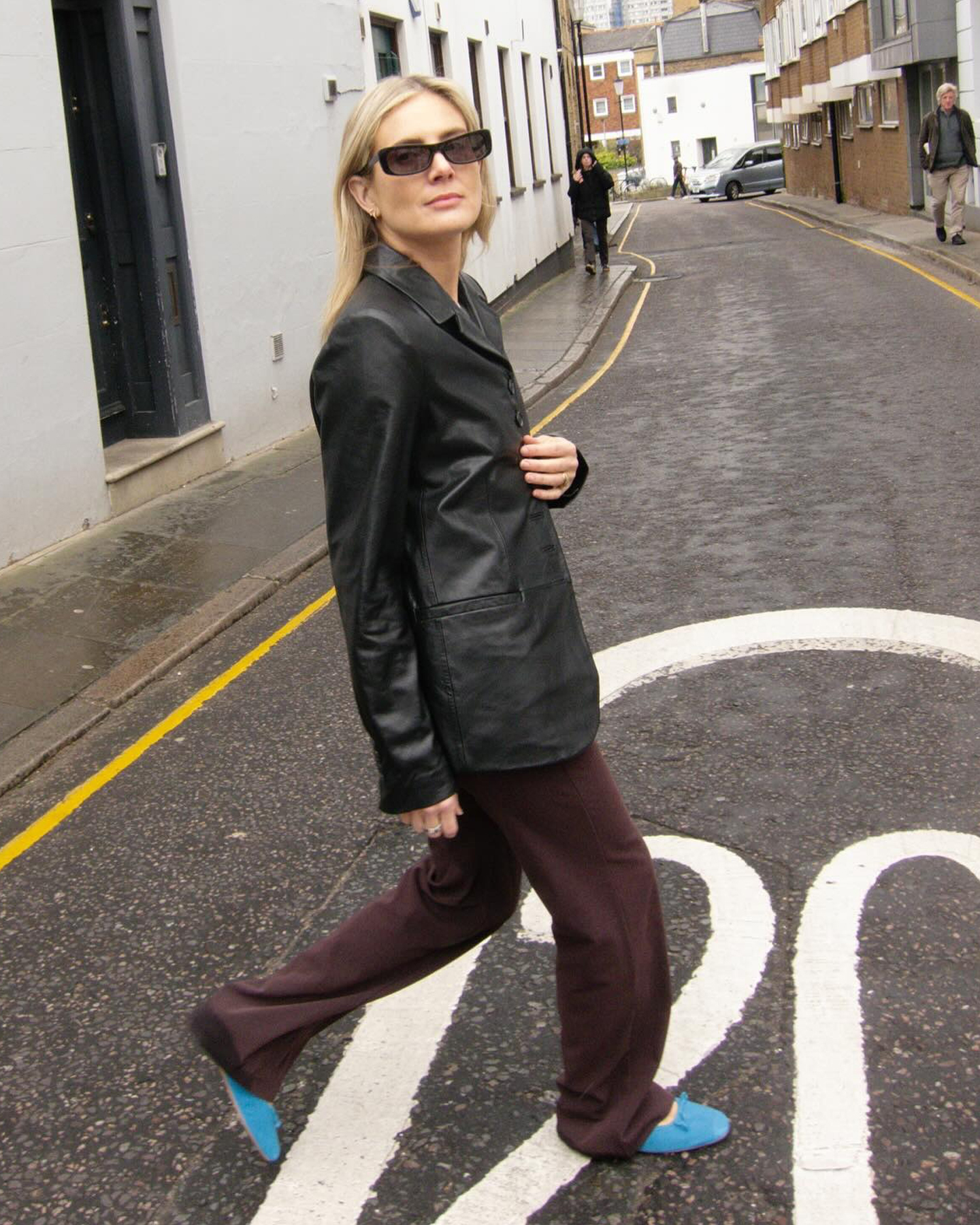 British style influencers Lucy Williams crosses a street in London wearing a black leather jacket, relaxed brown pants, and bright-blue ballet flats