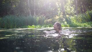 Woman laughing and swimming in pond with green foliage around to beat the heat