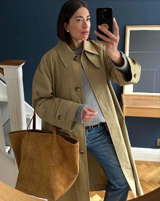 British influencer Lucy Alston poses for a mirror selfie wearing a collared trench coat, gray crewneck sweater, tan suede tote bag, black belt, and jeans