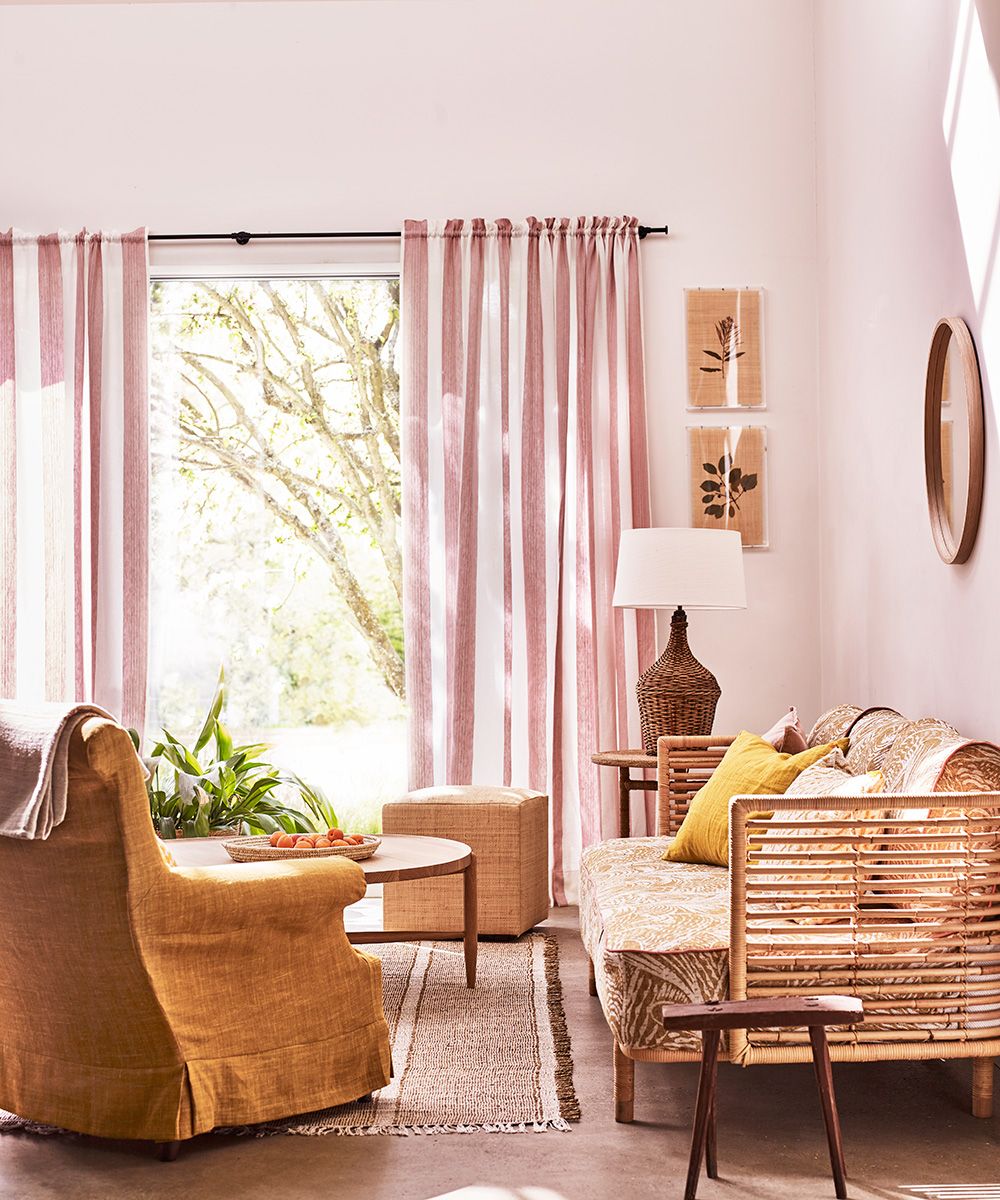 A summer living room with woven furniture, pink walls and pink striped drapes.
