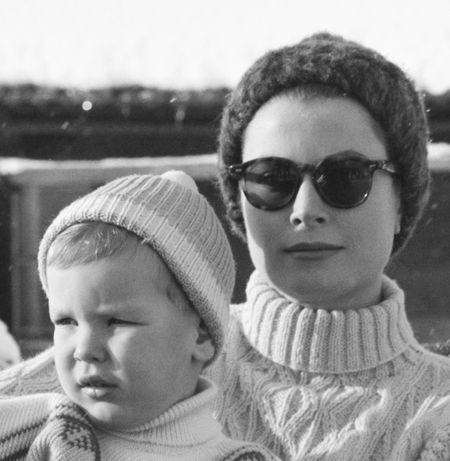 Grace Kelly holding young Prince Albert wearing sweaters and winter hats in front of a snowy tree