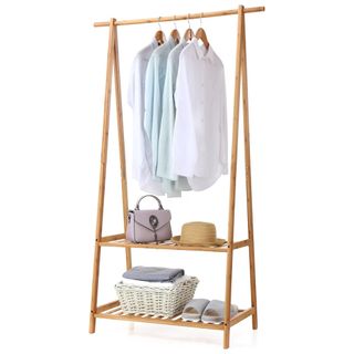 A wooden bamboo clothing rack with shirts hanging up, a shelf with a handbag and hat, with a white basket and slippers