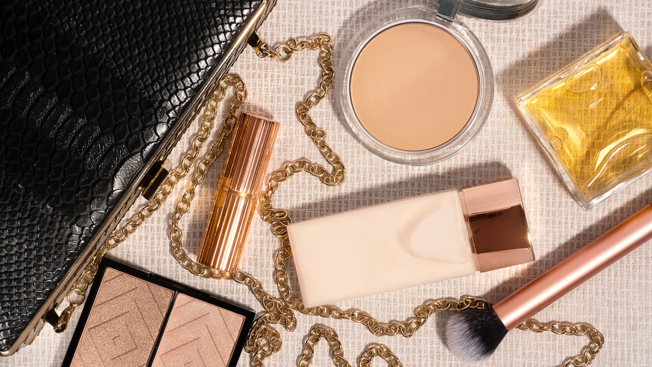 Bird&#039;s eye view of a black clutch bag, four make-up products and a brush and a bottle of perfume laid out in a flatlay against a cream background