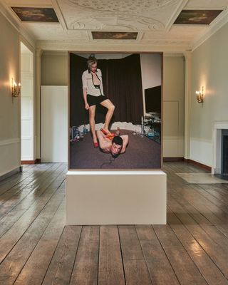 An installation view of an image featuring a naked male on the floor face down looking up with a woman wearing a white blouse, black skirt and red shoes with one foot on him.