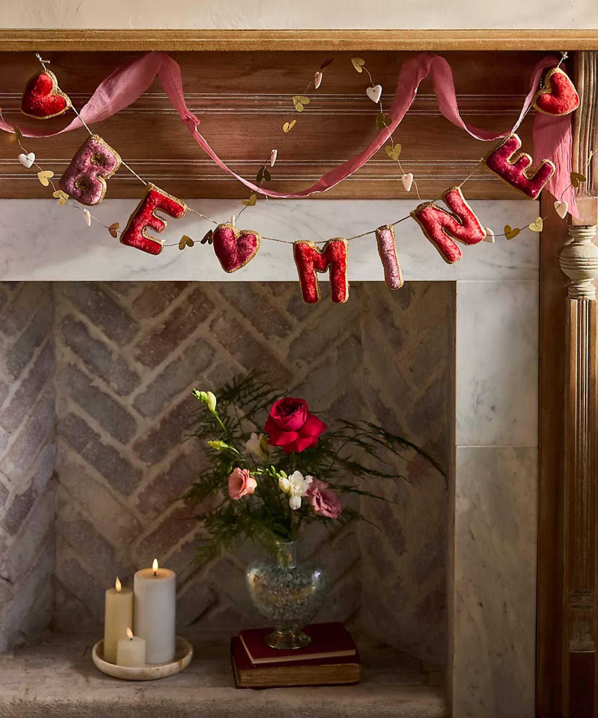 an empty fireplace with an assortment of candles and a vase of flowers inside, valentine's day inspired garland draped across the mantel