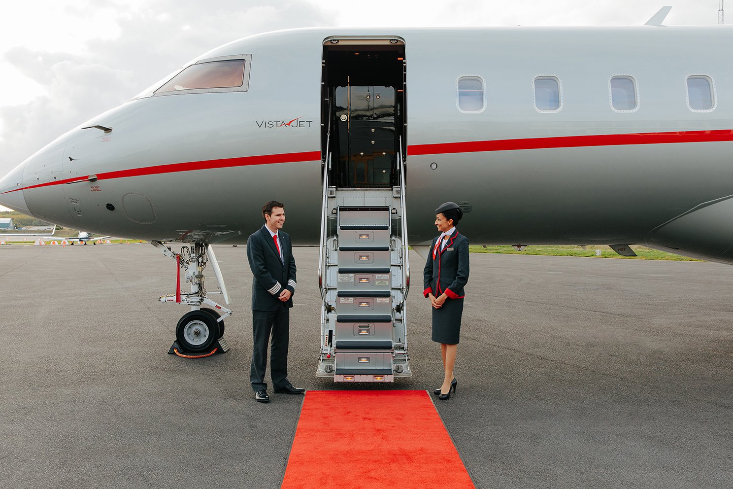 Two cabin crew stand at the foot of the steps up one of VistaJet jets, the staircase is reached by walking along a red carpet.