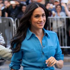 Meghan Markle smiling and wearing a blue shirt dress walking outside