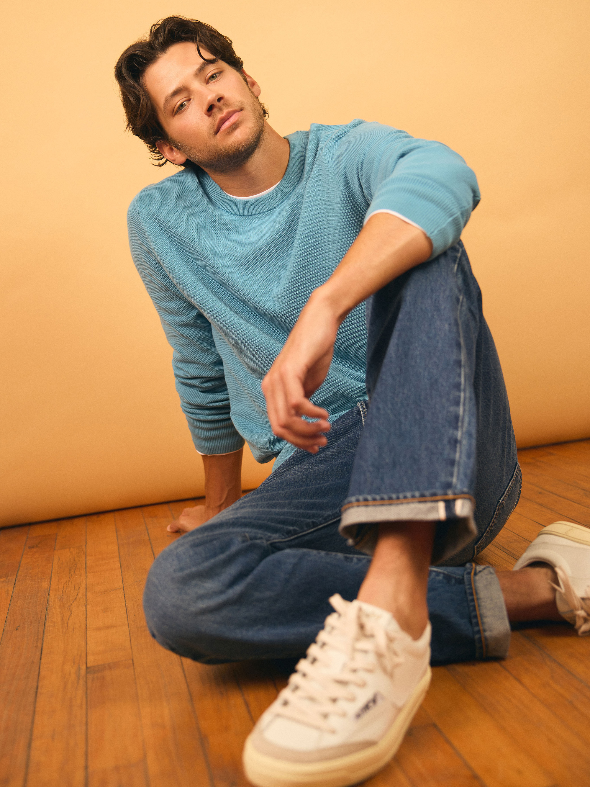 Matt Cornett photographed by Henry Wu and styled by Ashley Weston. He is sitting on a wood floor with his arm resting on his knee wearing a blue crewneck sweater, jeans, and white sneakers.