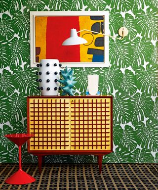 Colorful hallway with plant print green wallpaper, red and yellow cabinet, glossy red stool, colorful abstract artwork, white and metal wall lamp, cabinet decorated with decorative vases and ornaments, black and brown patterned carpet