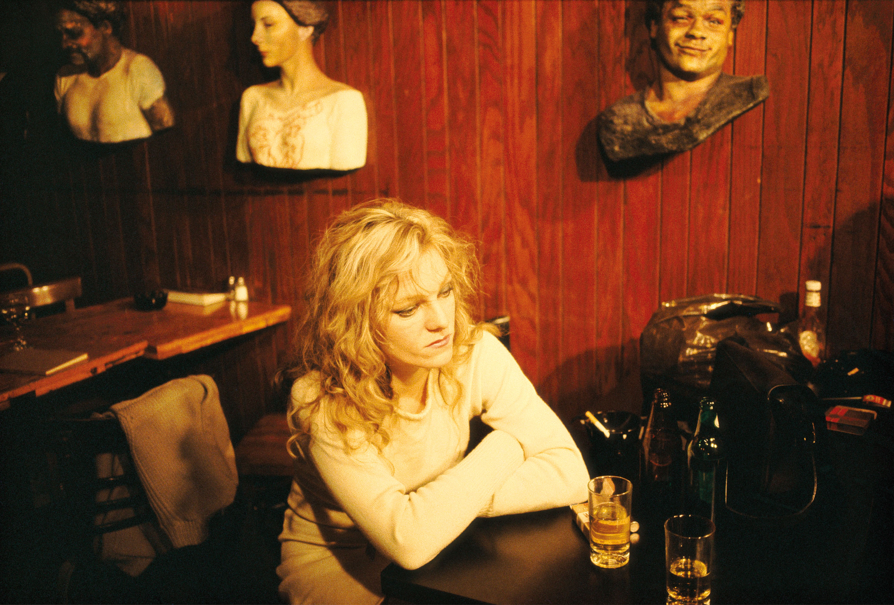 A blonde woman sits alone at a dark bar table with drinks and cigarettes, looking downward in front of a wood-paneled wall featuring sculpted busts.