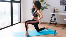 woman performing a forward lunge on a turquoise exercise mat in a home office setting with a large window in front of her and white desk behind her.
